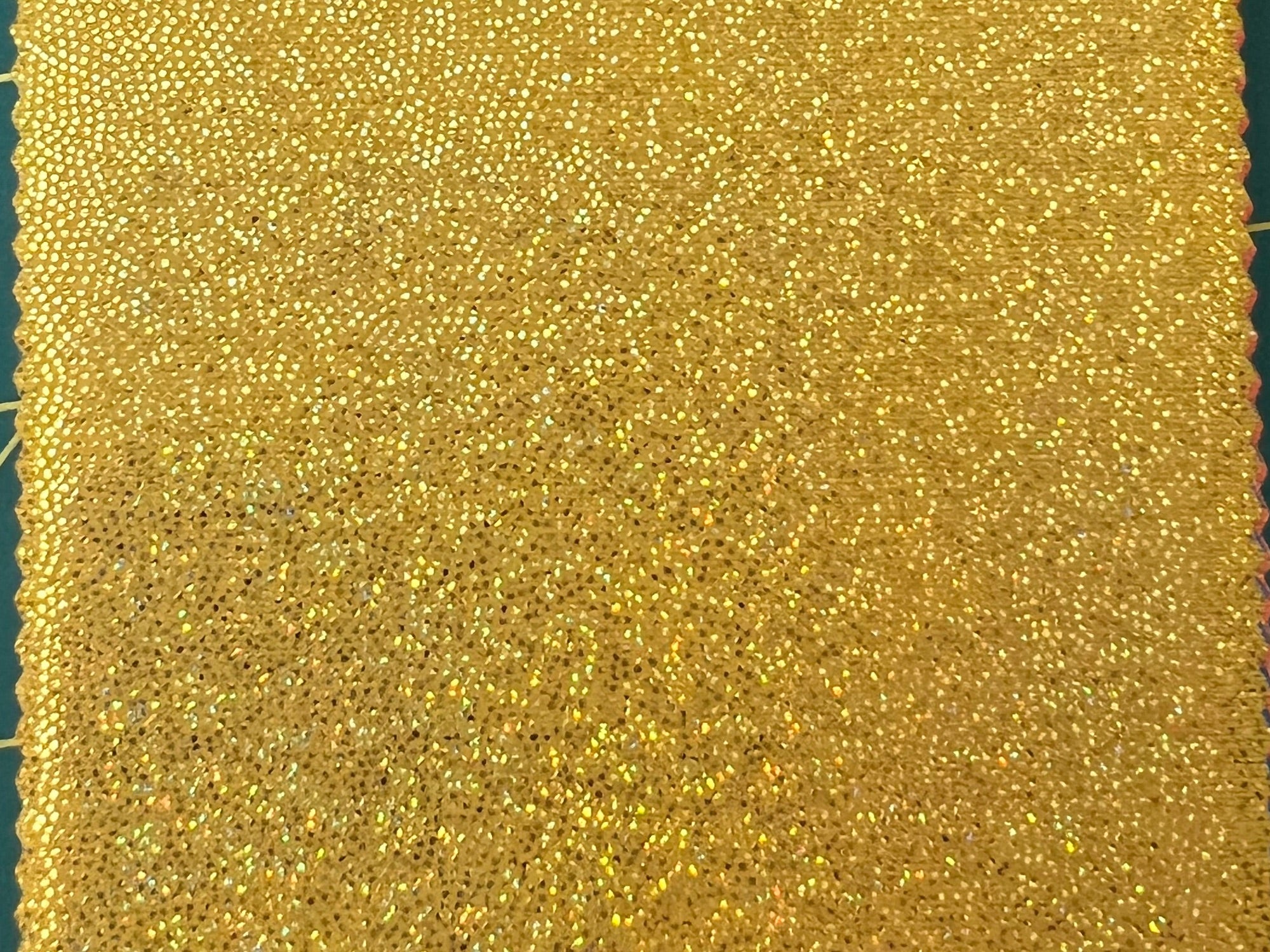 A close-up of a gold sequined fabric texture with shimmering metallic highlights and scalloped edges visible on the left and right sides.