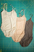 Three swimsuits in beige, pink, and brown on a grid-patterned surface.
